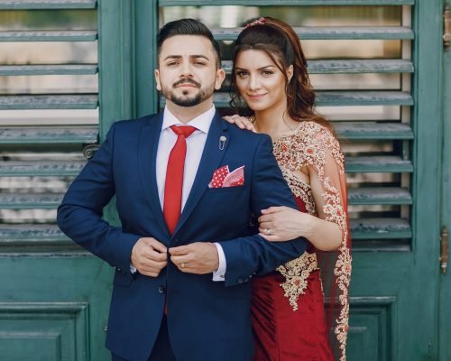 Beautiful woman in a long red dress walks around the city with her husband in a blue suit and with a beard